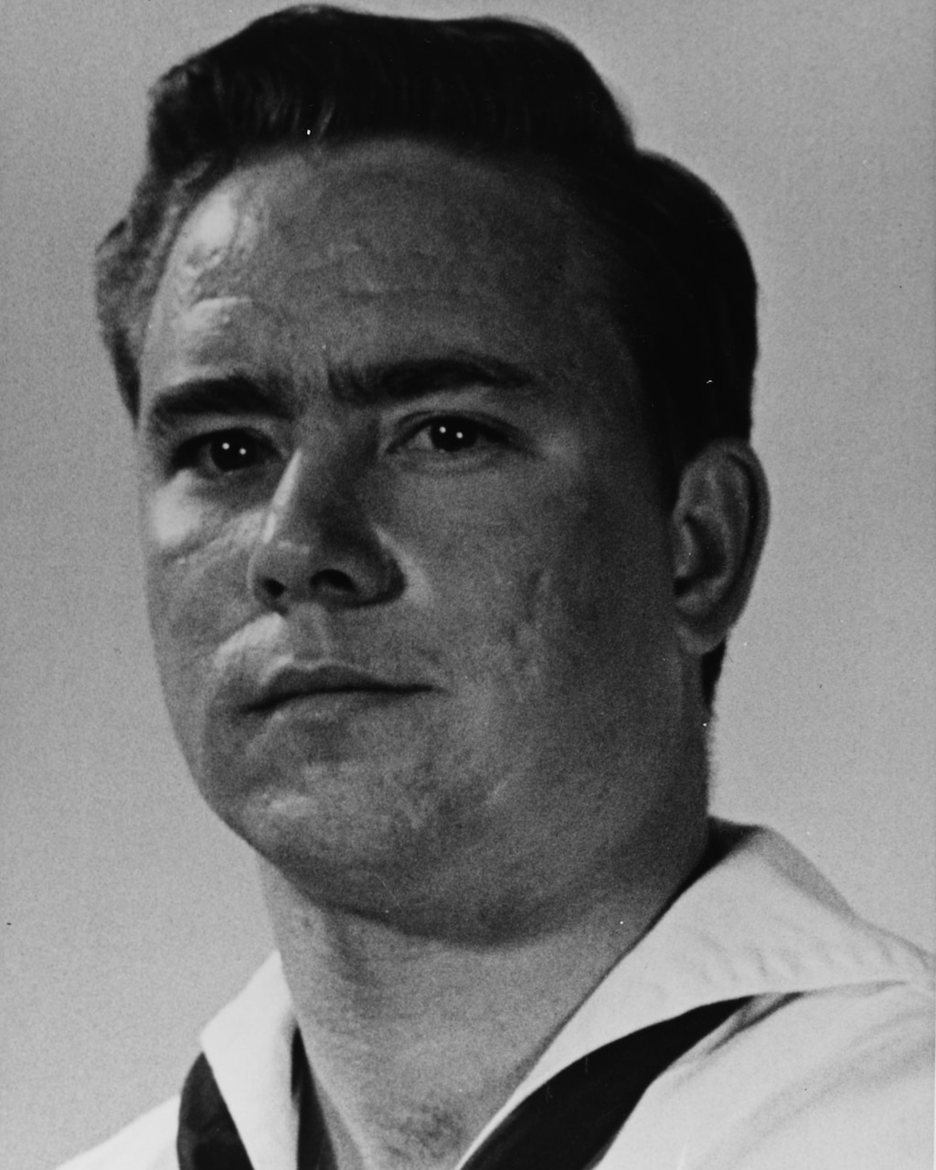 A sailor poses for a black-and-white photo in his dress white Navy uniform.