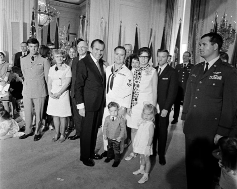 A sailor poses for a photo with a woman, a man and two small children amid a crowd of people.