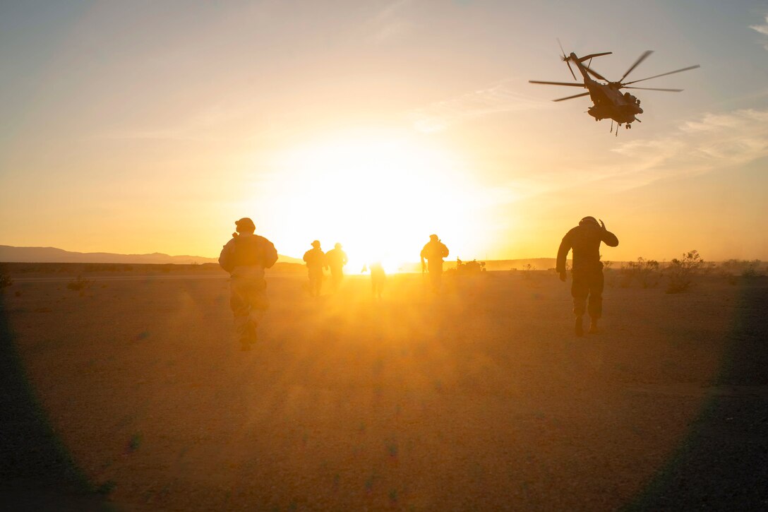 Marines run in a field as a helicopter flies above under a sunlit sky.