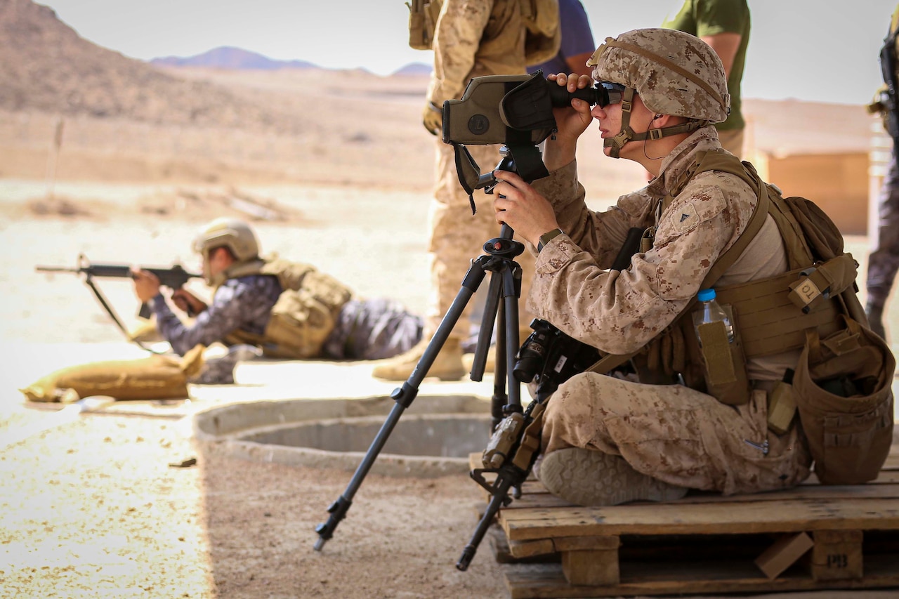 A Marine looks through an eyepiece, as another service member appears to fire a weapon.