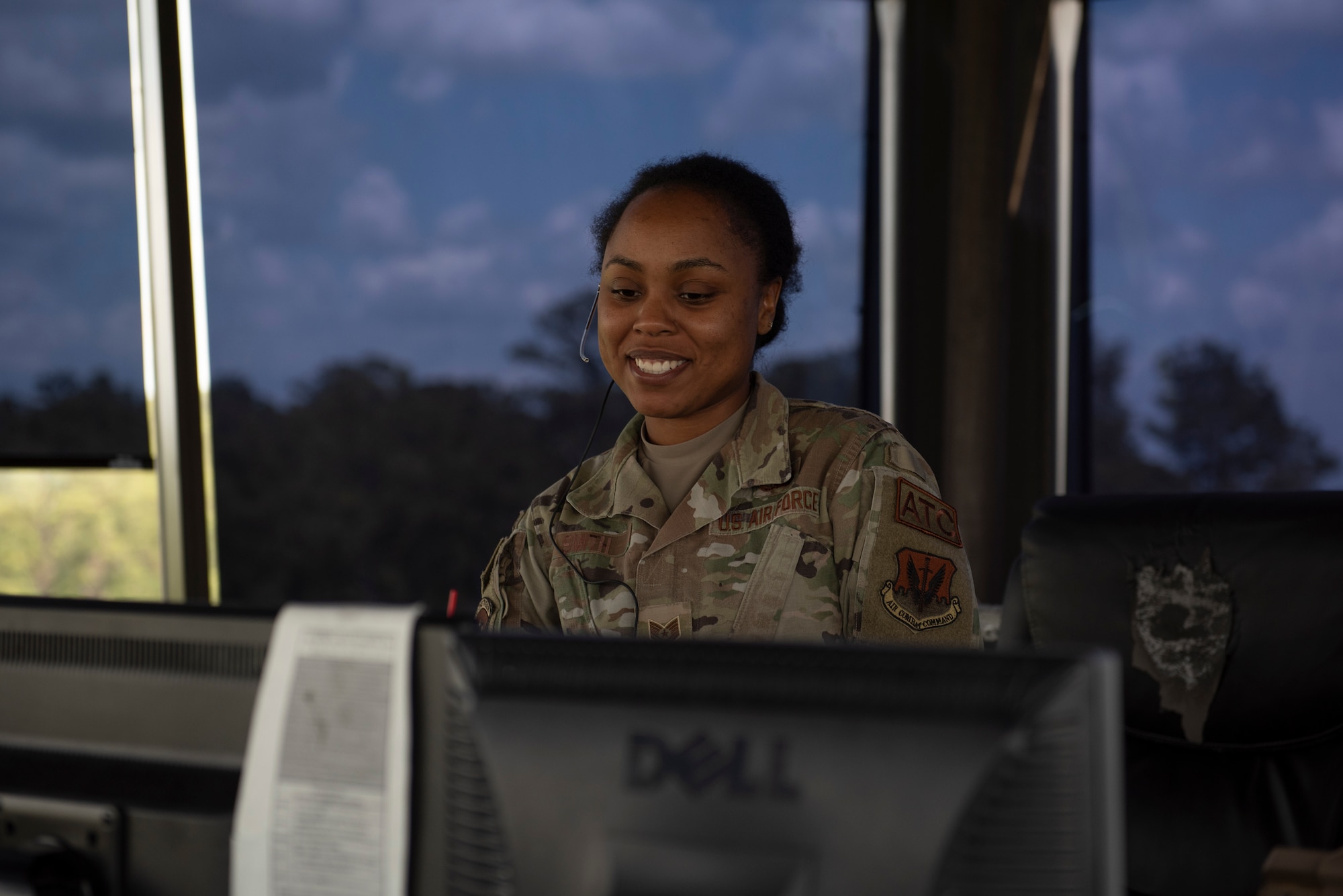 A photo of a woman smiling, standing in front of a computer.