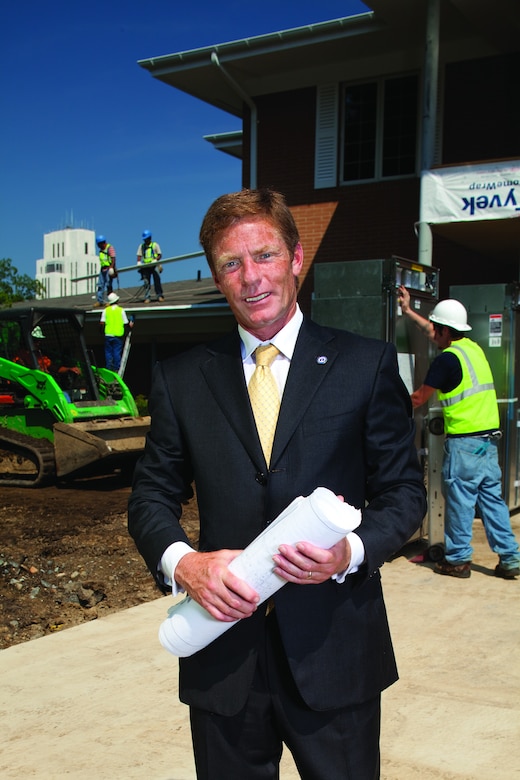A man wearing a business suit holds rolled up papers. A construction site is in the background.