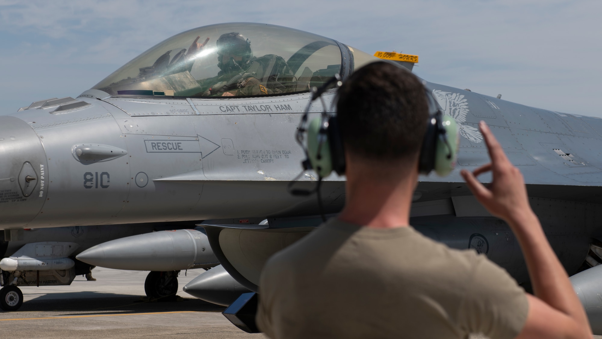 A fighter jet pilot waves at a ground crew airman from a fighter jet cockpit.