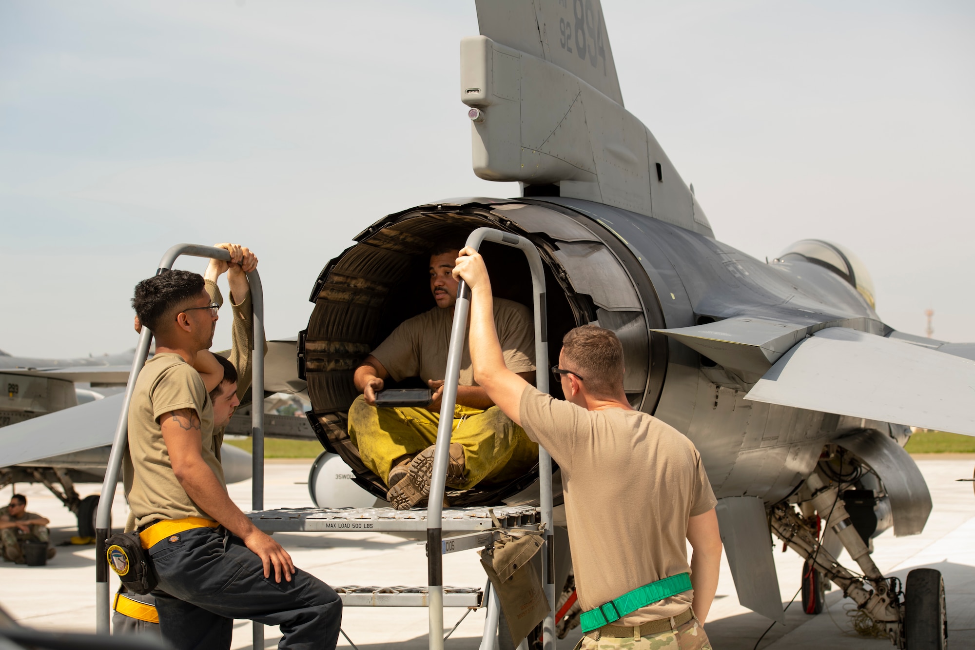 An Airman sitting in the engine at the rear of an F-16C Fighting Falcon and flanked by three other Airmen.