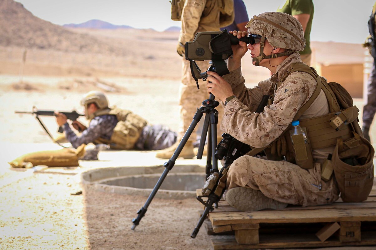 A Marine looks through an eyepiece, as another service member appears to fire a weapon.