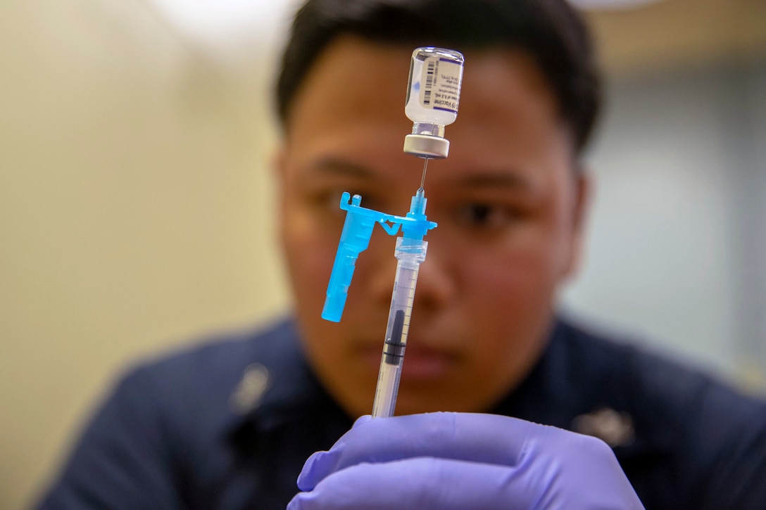 A sailor wearing gloves holds a syringe that’s been inserted into a small bottle.