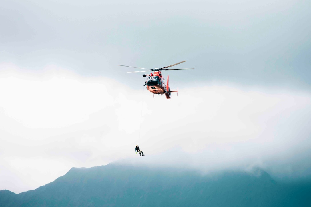 A Coast Guardsman hangs from an airborne helicopter by a rope.