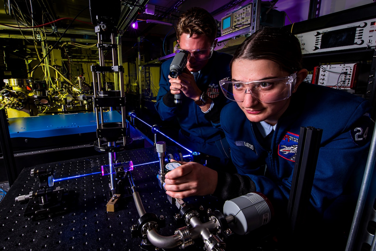 Two young people in uniform manipulate a scientific apparatus that uses lasers.