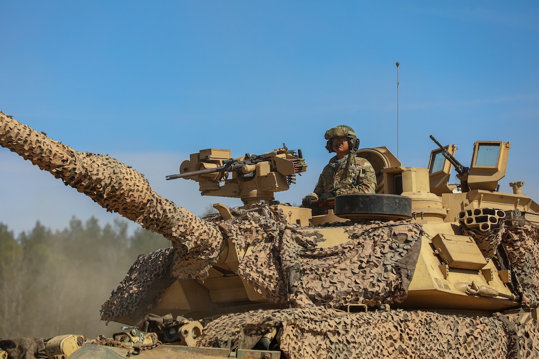 A soldier stands in behind a mounted machine gun and in a hatch of a camouflage netting-covered tank.