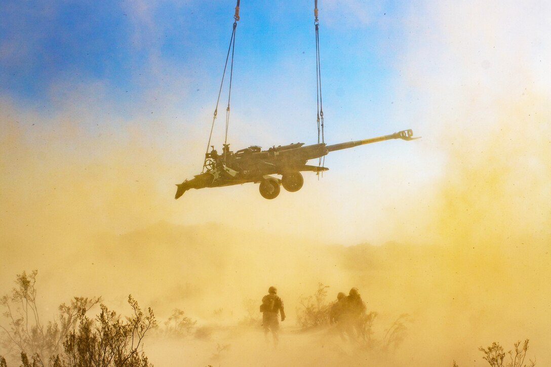 A howitzer is lifted airborne amid waves of dust while Marines watch.