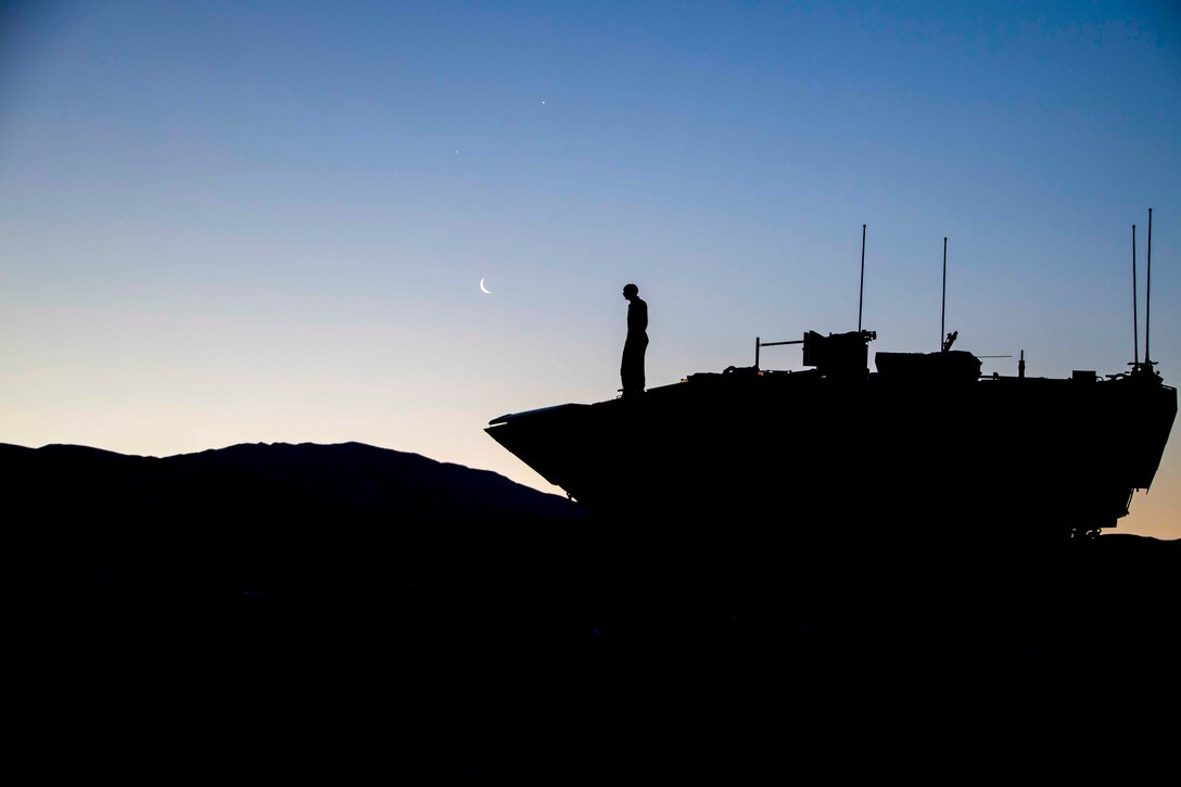A Marine shown in silhouette stands on top of a tank.
