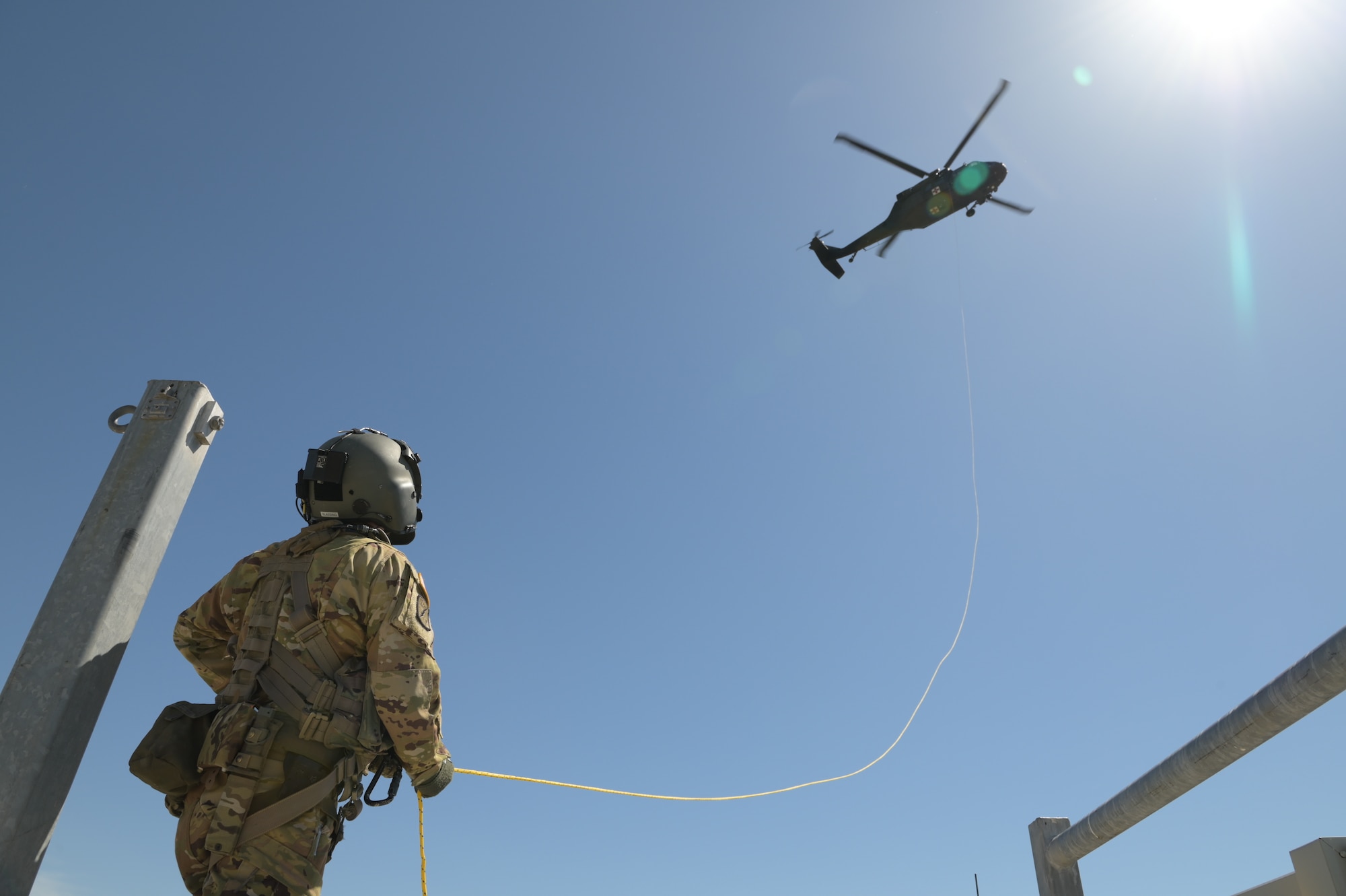 An Army Soldier is standing on the ground tethered to a UH-60 Blackhawk Rescue Helicopter