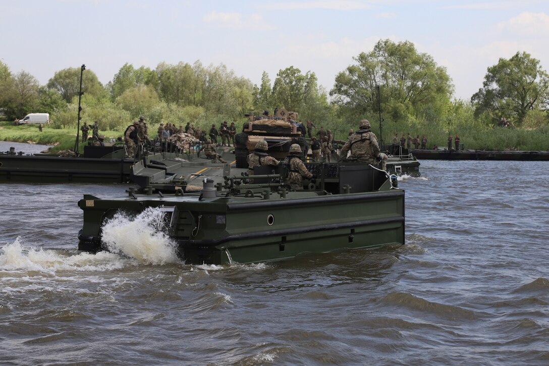 Soldiers drive a vehicle across a river while more soldiers and a tank sit atop a floating bridge in the background.