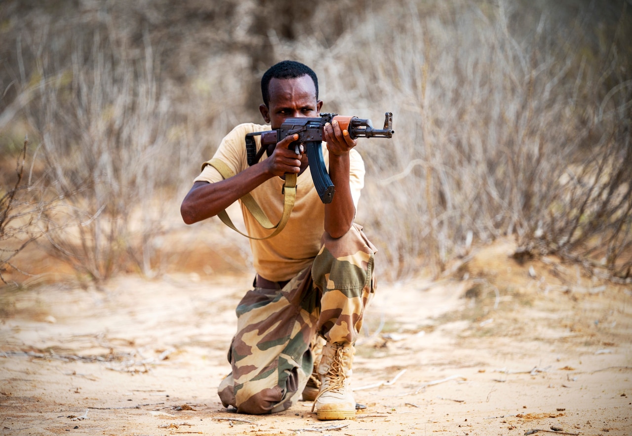 A man in a military uniform kneels on one knee and aims a rifle.