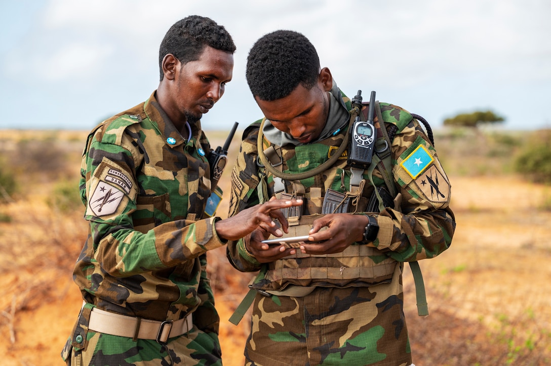 Two men in military uniforms stand near each other outdoors and look at a cell phone.