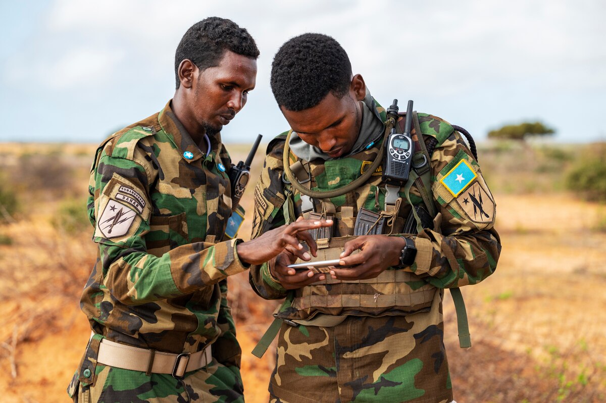 Two men in military uniforms stand near each other outdoors and look at a cell phone.