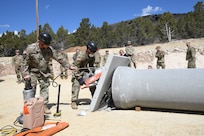 A soldier uses a hose to spray water on a concrete saw being used by another soldier cutting through a concrete barrier.