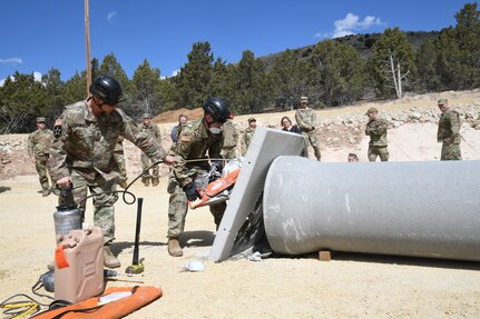 A soldier uses a hose to spray water on a concrete saw being used by another soldier cutting through a concrete barrier.