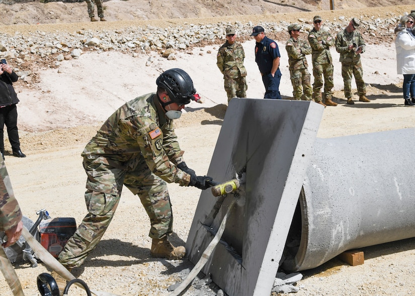 A soldier uses a sledge hammer to pond and opening in a concrete barrier