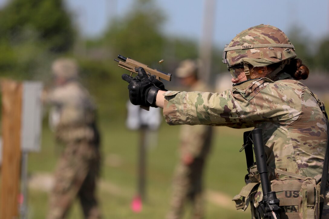 A soldier fires a weapon as fellow service members stand in the background.