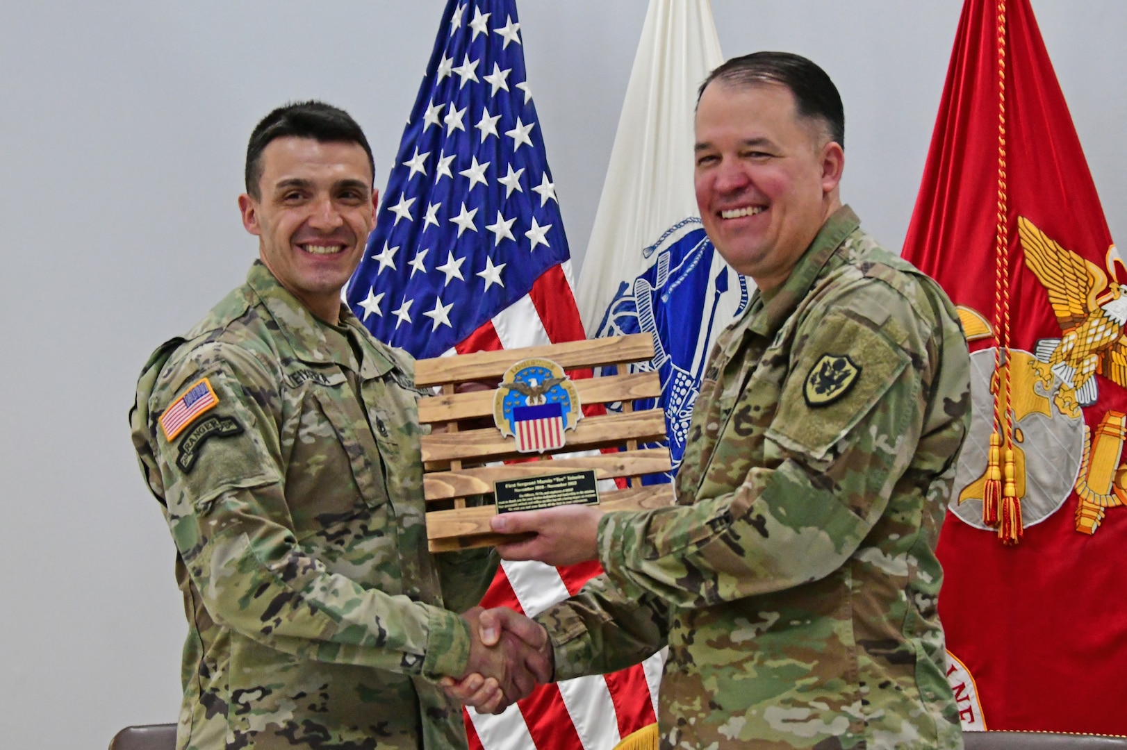 Two men in uniform exchange an award honoring one of them for their service. The award is shaped as a pallet.