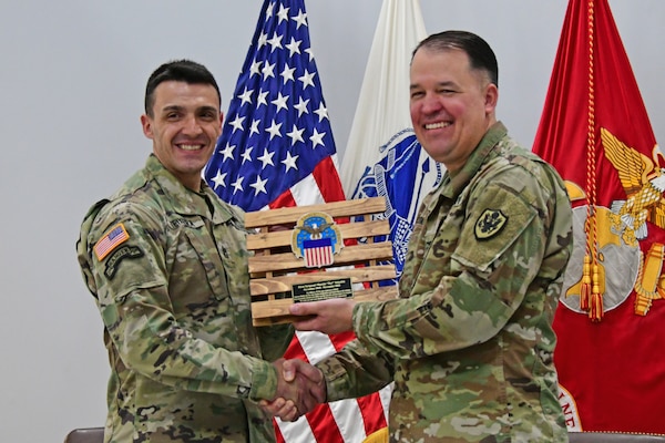 Two men in uniform exchange an award honoring one of them for their service. The award is shaped as a pallet.