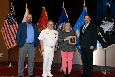 Woman holding an award posing with three men, in front of U.S. flags.