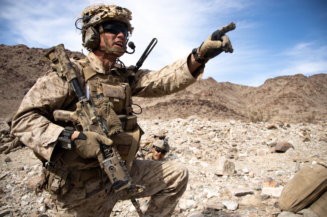 A Marine carrying a weapon and gear, points in a rocky, desert area.