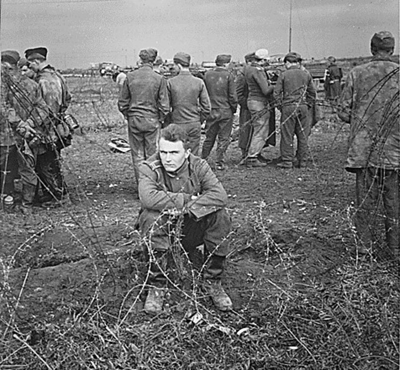 A man sits with his arms crossed near barbed wire while several others stand around behind him.