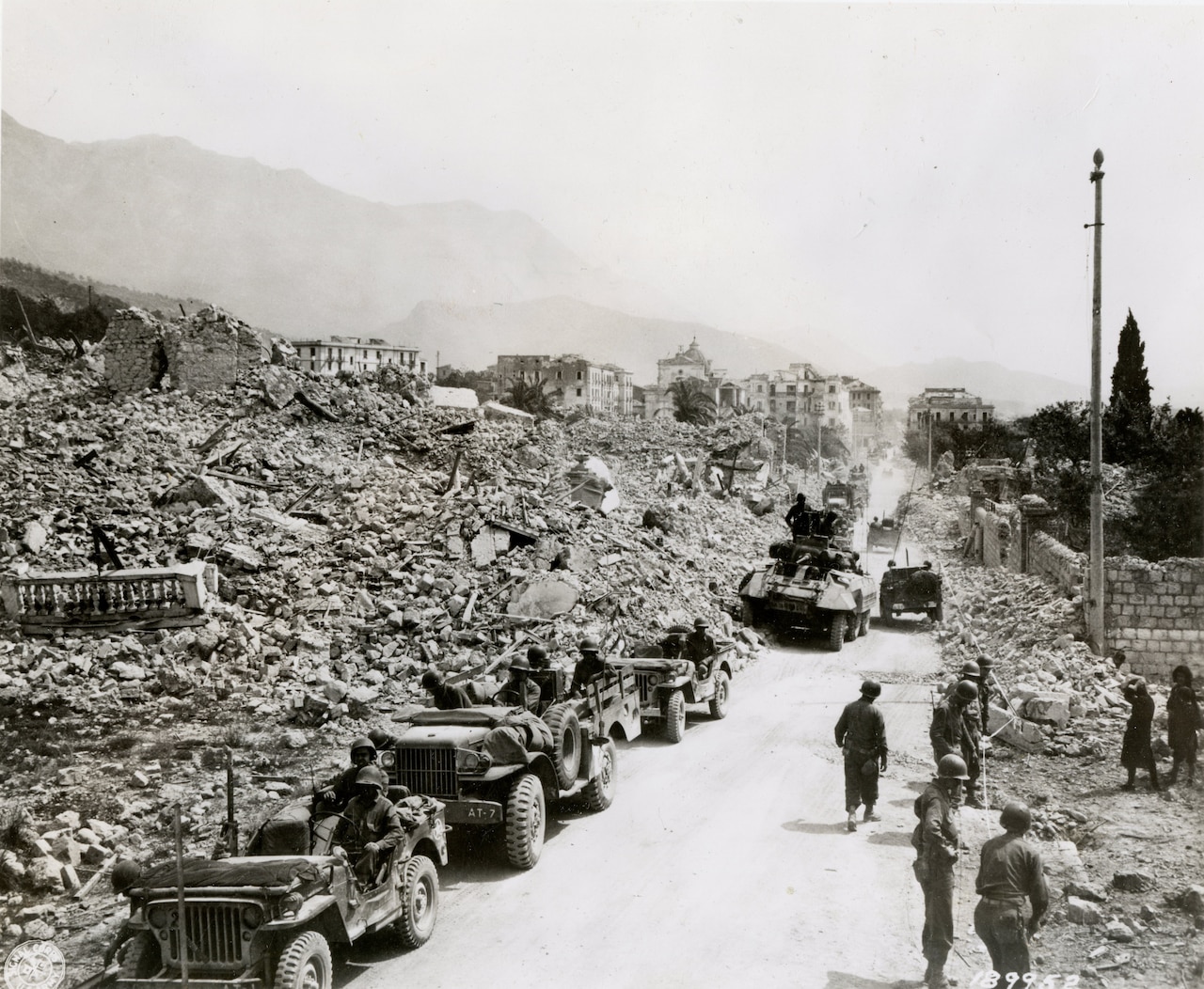 Tanks and soldiers transit a road that is partially obstructed by rubble.
