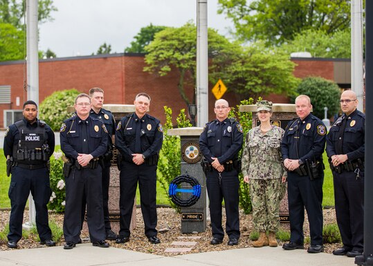 Several police officers and a woman in US Navy utility uniform with a black and blue wreath in front of a memorial. 