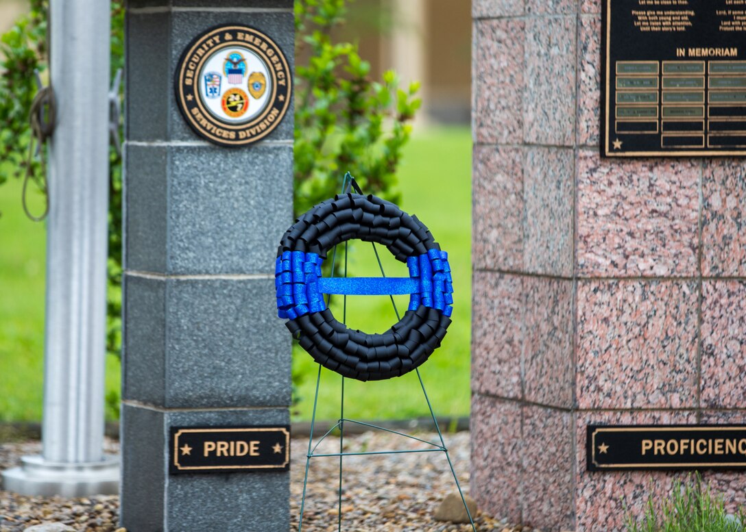 A black wreath with a blue line sits in front of a memorial outside.