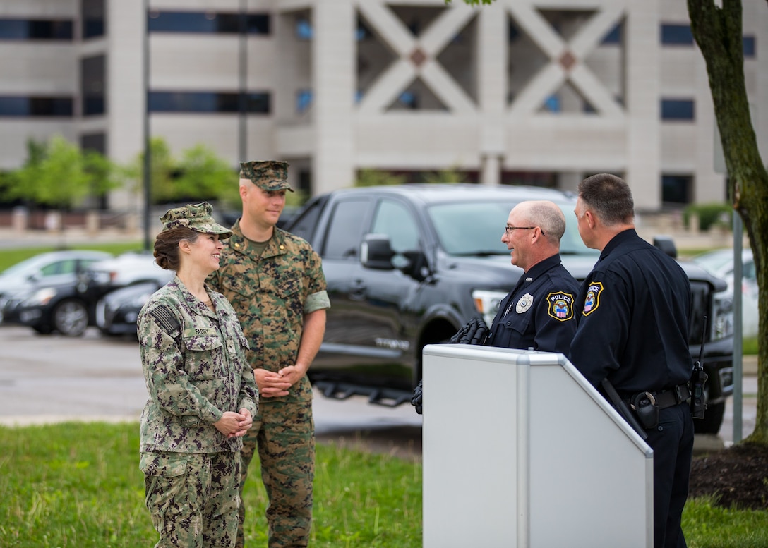 One man in a US Marines uniform stands next to a woman in a US Navy utility uniform speaking to two police officers in front of a outdoor podium.
