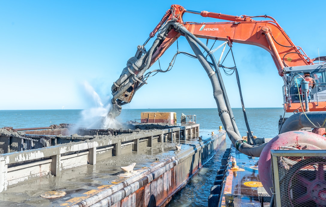 An orange excavator on a barge pumps water into a scow holding dredged sediment.