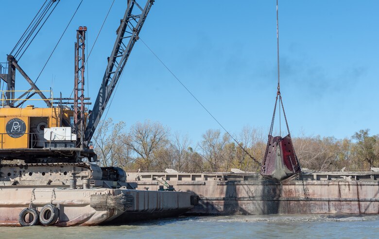 A yellow crane on a barge with a red clamshell bucket scoops sediment from a waterway.