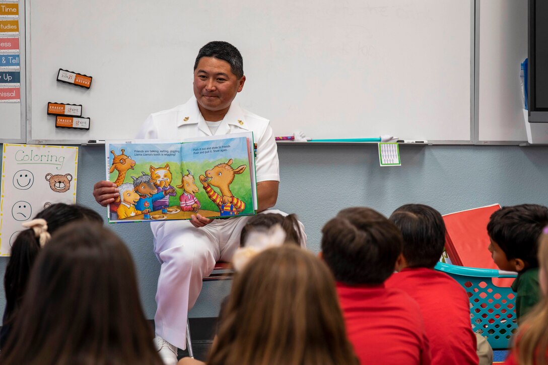 A sailor holds open a book for small children in a classroom.