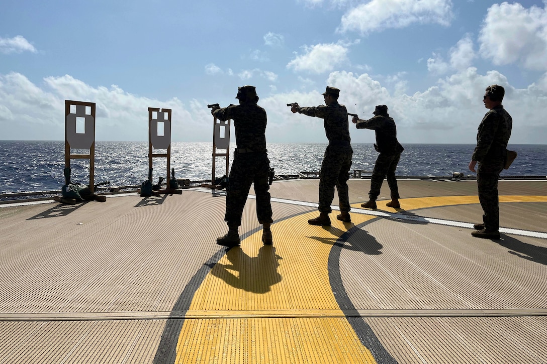 Three Marines fire at targets as a fellow service member watches aboard a ship at sea.