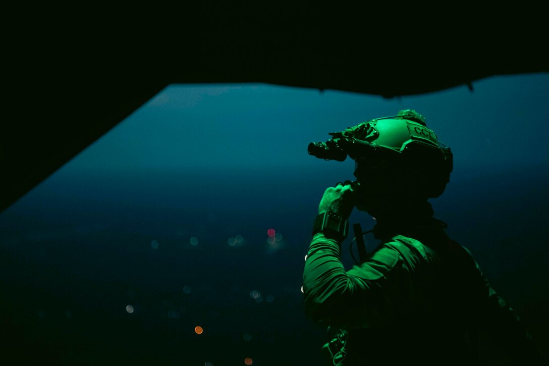 Green light shines on an airman standing inside an airborne aircraft at night.