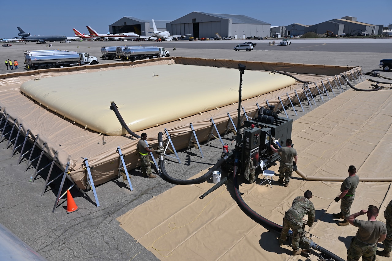 Several uniformed personnel stand near large hoses and pumps that are feeding into a large, flat, inflated container on an airfield.