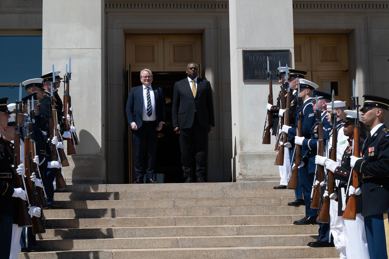 Two men stand side by side at the top of a stairway outside of a building; service members form an honor guard on either side of the stairs.