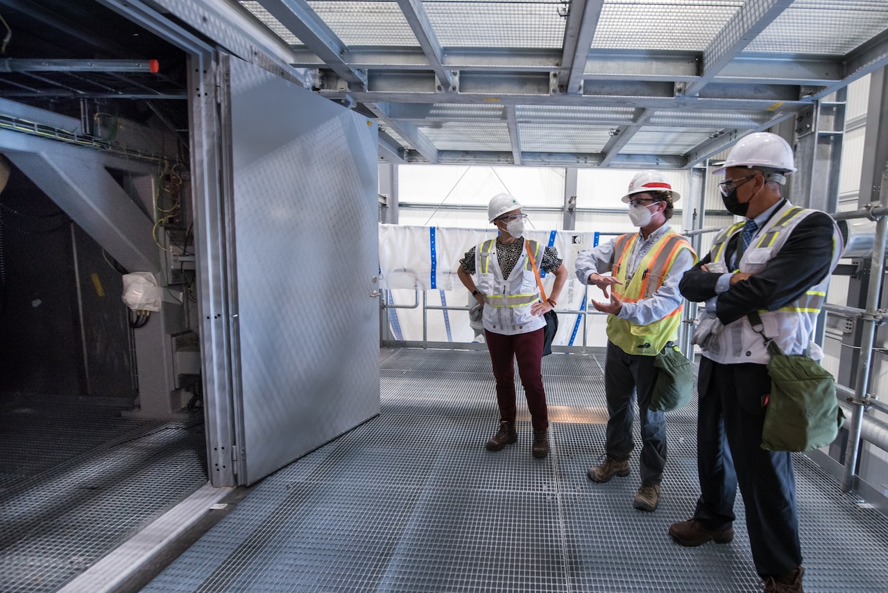 Three people wearing hard hats and masks stand next to one another inside a plant.