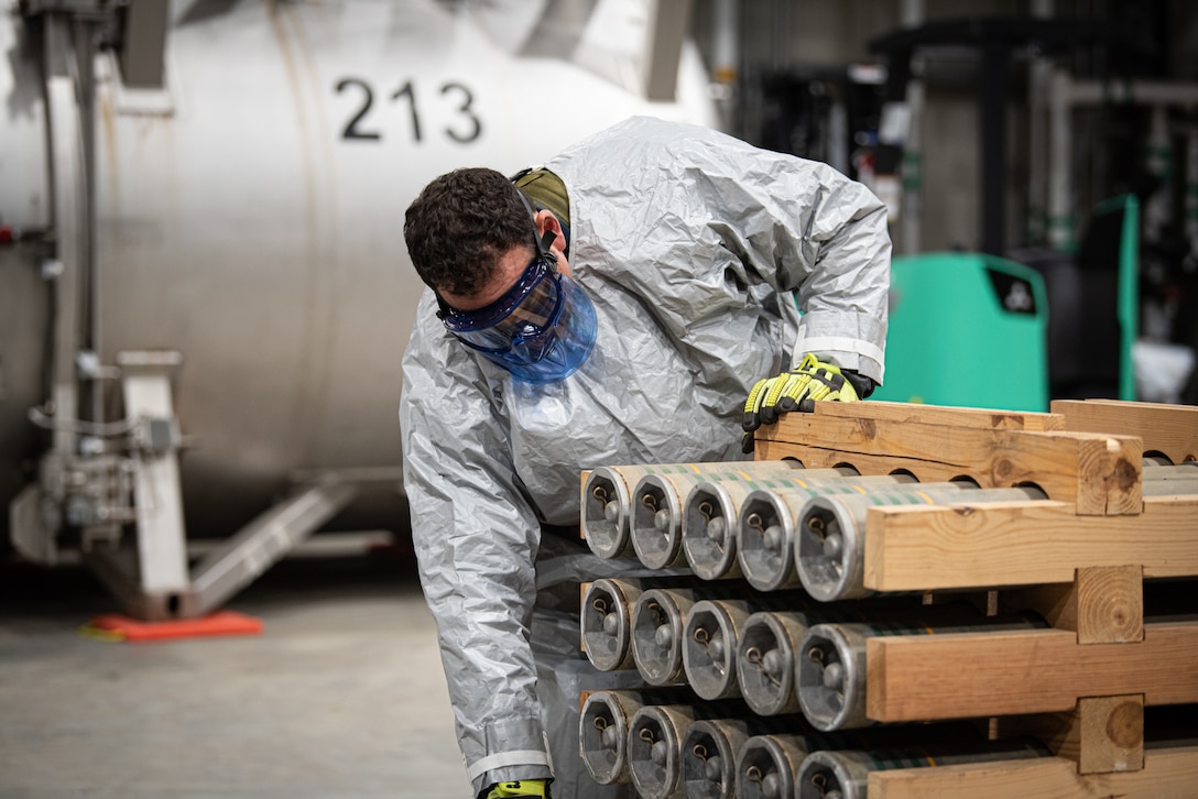 A man wearing protective gear looks at metal canisters inside a wooden grate.