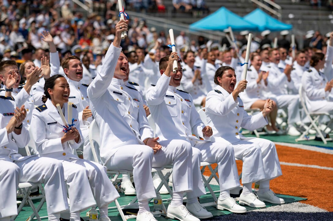 A group of Coast Guard graduates in white cheer while seated.