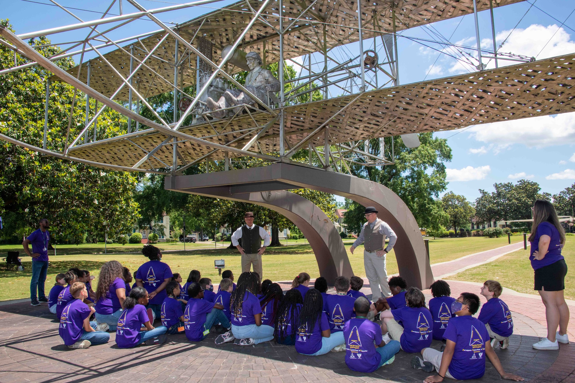 Maxwell AFB, Ala. – Elementary students from Bear Exploration Center For Mathematics, Science and Technology School perch in the shade of the Wright Flyer replica as they learn about the history of aviation from “Orville” and “Wilbur Wright,” May 13, 2022. (US Air Force photo by Melanie Rodgers Cox/Released)