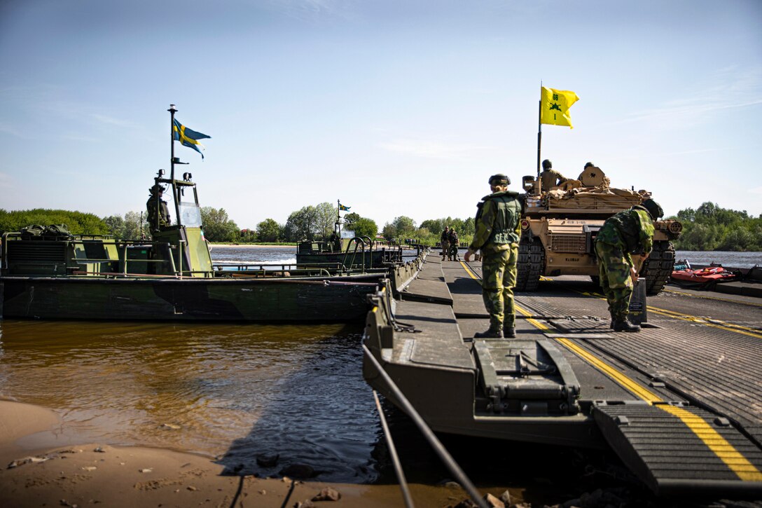 Soldiers and NATO allies transport a tank across a body of water.