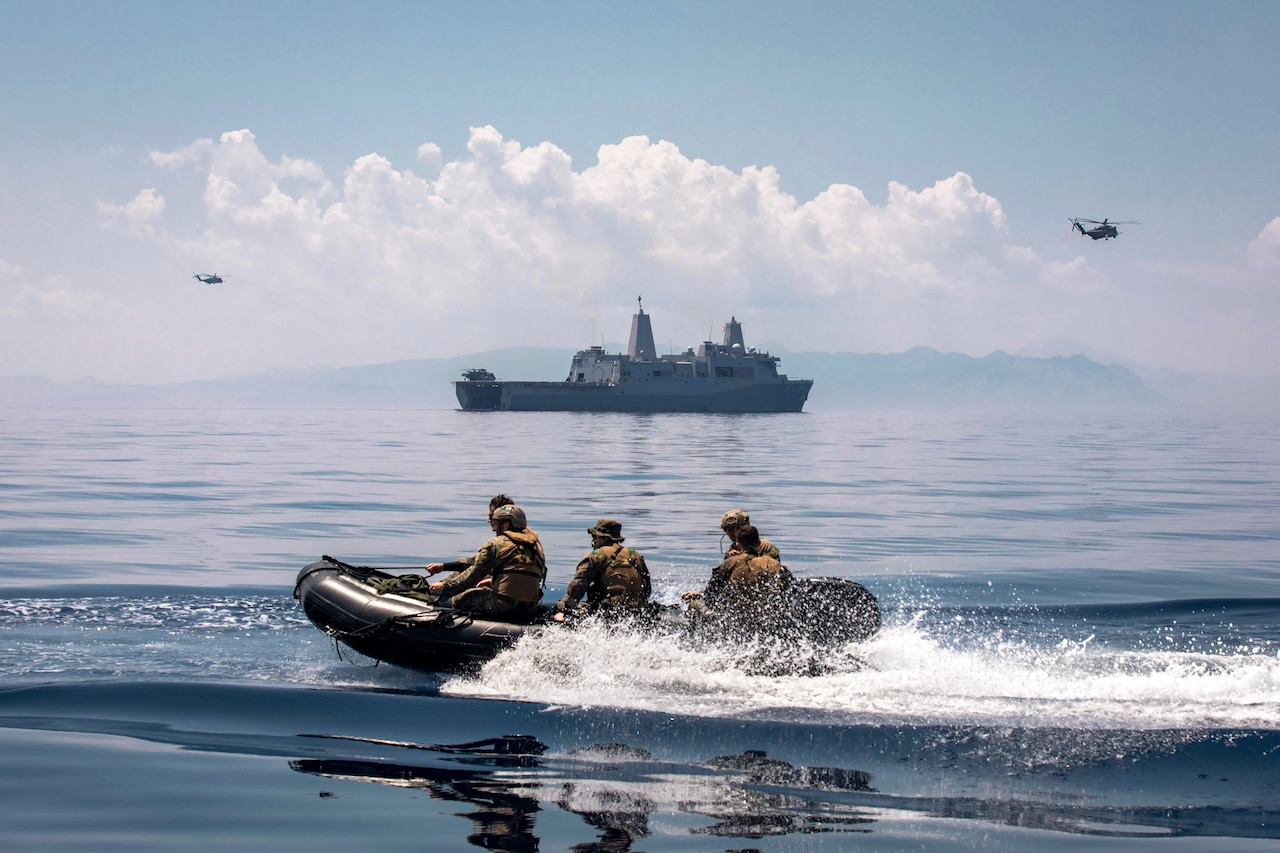 Marines ride in a small with near a large ship and two hovering helicopters.