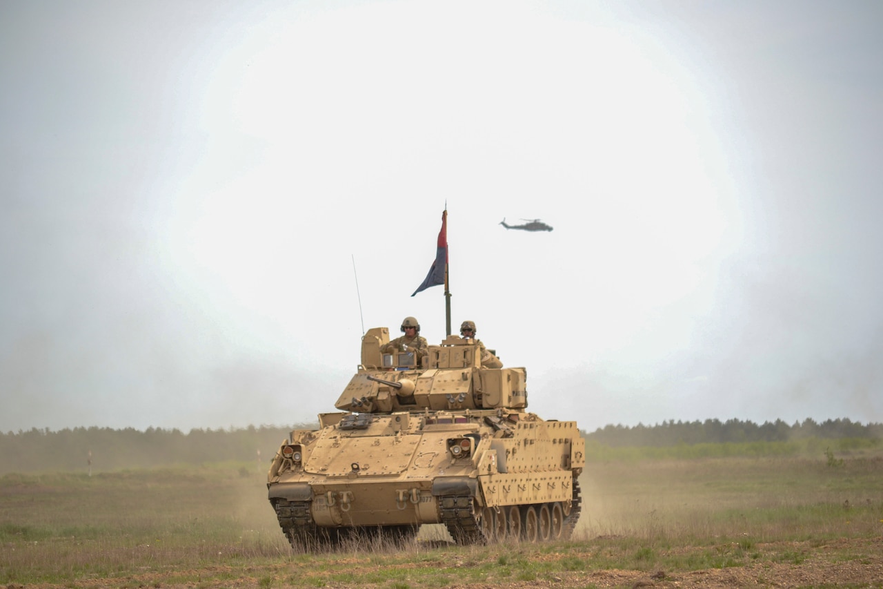 Soldiers stand in a tank while a helicopter flies above them.