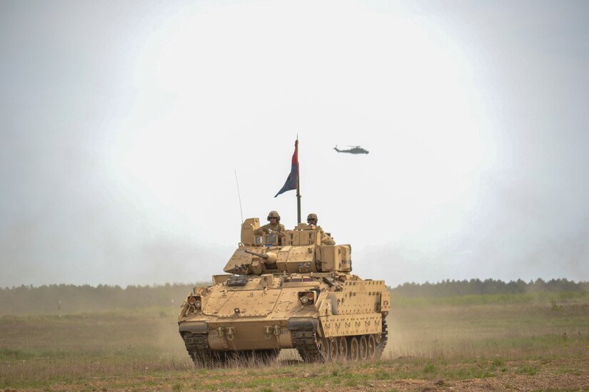 Soldiers stand in a tank while a helicopter flies above them.