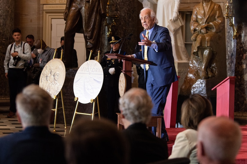 A man gestures broadly with his left arm while standing at a lectern.