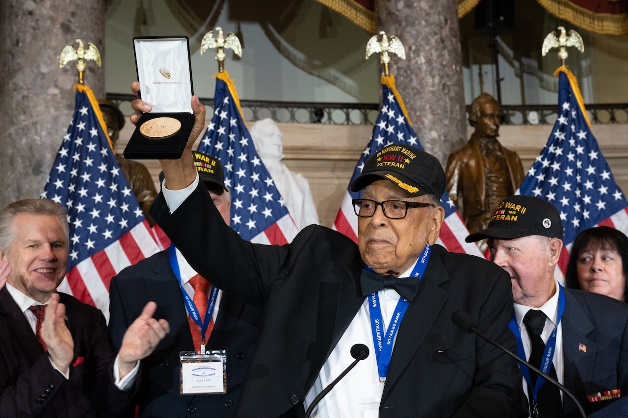 A man wearing a WWII veteran hat holds up a presentation case containing a large gold medal.